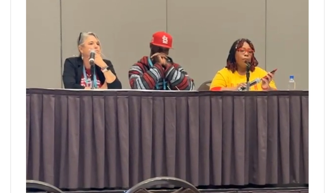 Three individuals seated at a table, each with a microphone, engaged in a discussion or interview setting.